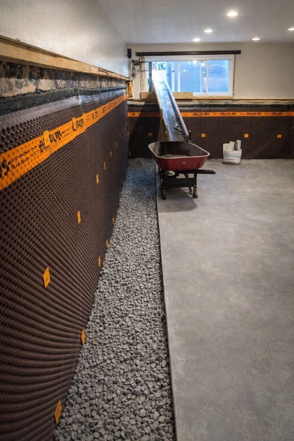 Rénovation d'un sous-sol montrant une barrière anti-humidité à picots sur les murs, une tranchée de drainage en gravier et une brouette près d'une fenêtre.