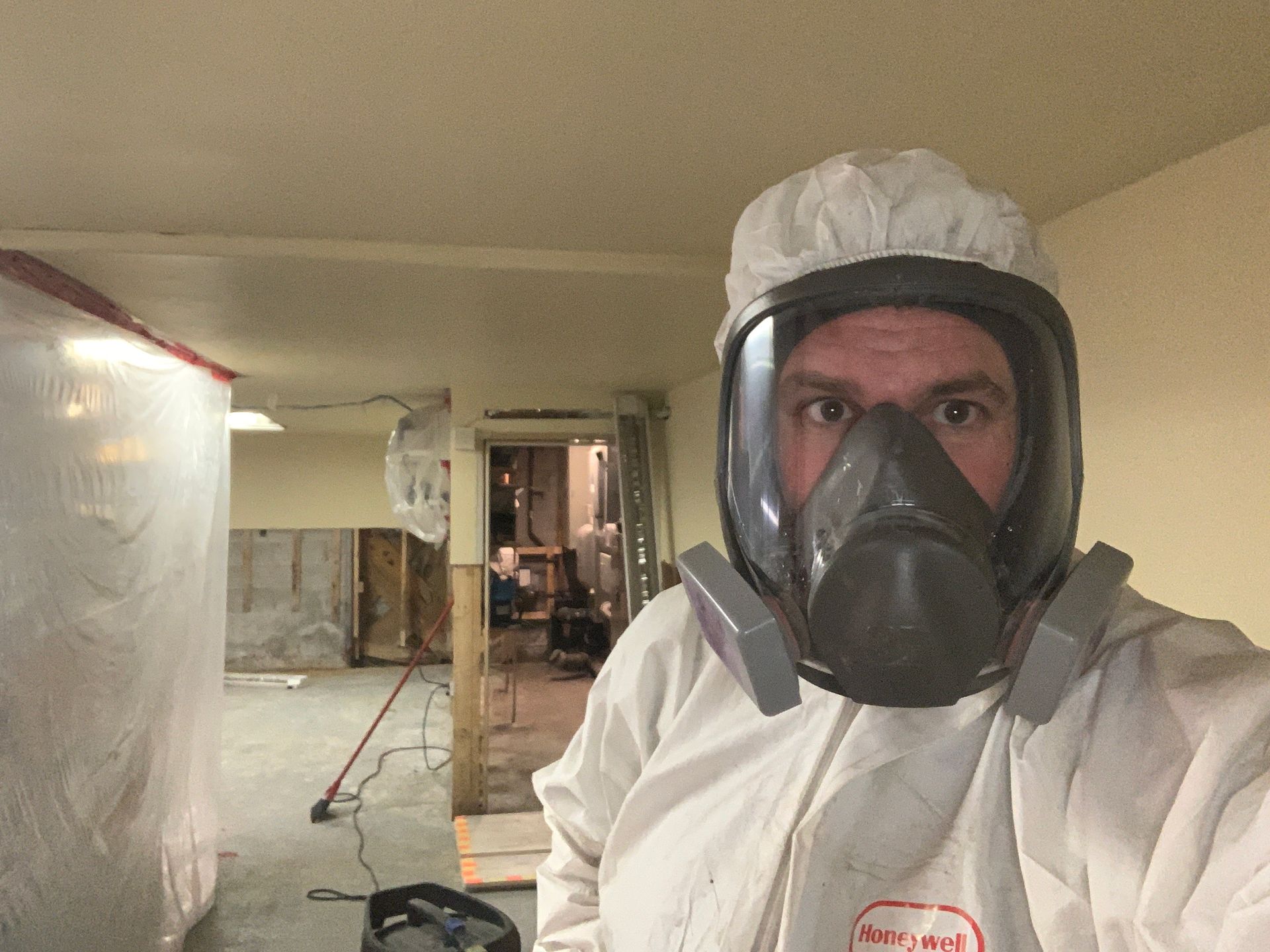 Une personne vêtue d'une combinaison de protection blanche et d'un masque respiratoire intégral se tient dans une pièce en cours de nettoyage après des travaux de construction.