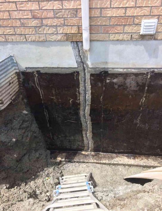 Une fissure verticale dans un mur de fondation en béton sombre et excavé est réparée avec un nouveau mastic, directement sous une descente de gouttière.
