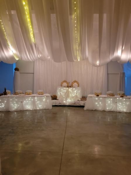 Decorated event hall with draped white ceiling fabric, string lights, and floral tables along a central aisle