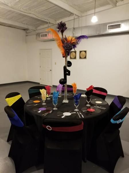 Round banquet table with black cloth, colorful place settings, and tall feather centerpieces in a white room