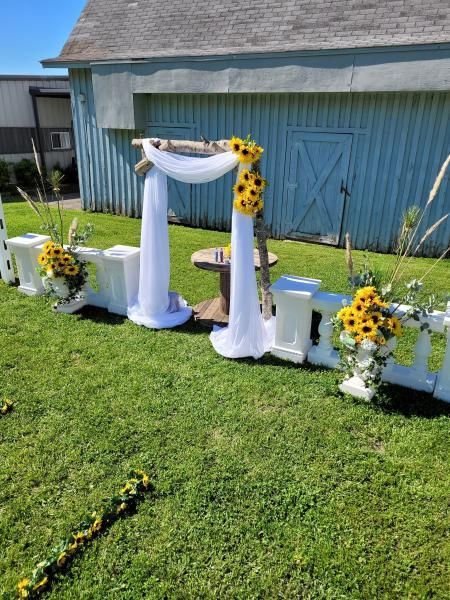 Outdoor wedding arch with white drapes, sunflowers, and blue accents on a grassy lawn
