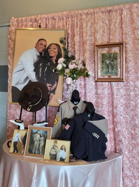 Memorial display with framed photos, flowers, hat, and black clothing on a table before pink curtains