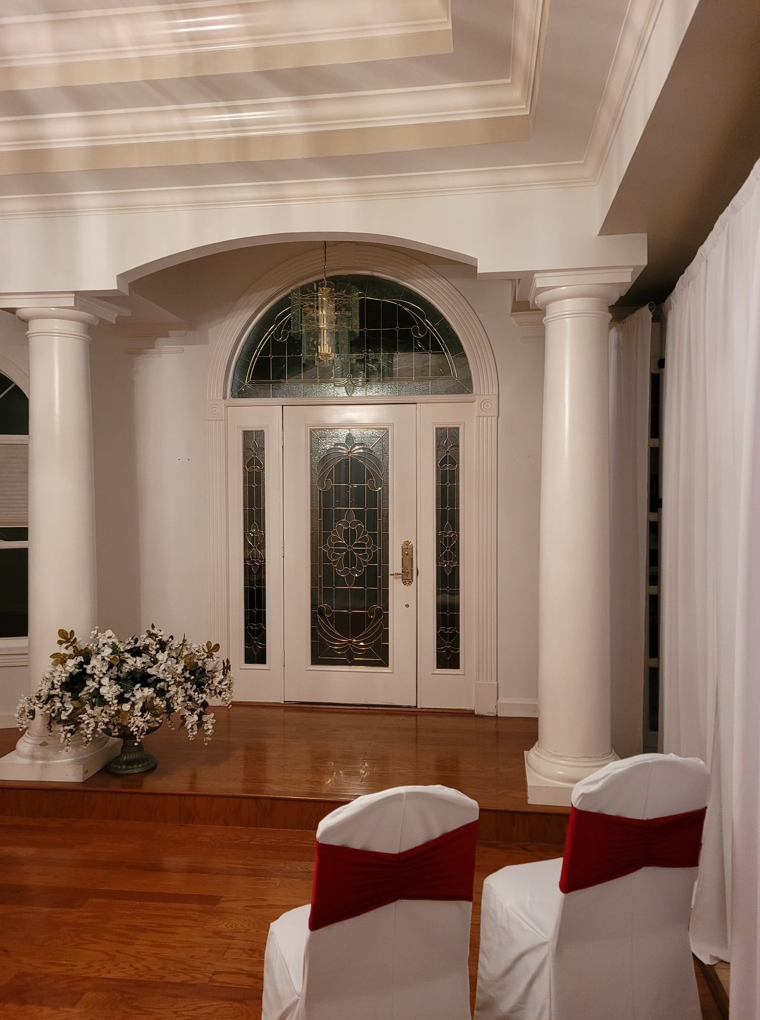 Elegant foyer with white columns, arched doorway, floral arrangement, and white chairs with red sashes on a wood floor