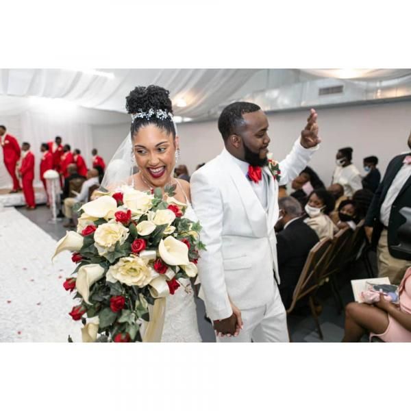 Bride smiling with a large bouquet at a wedding ceremony, while groom in white waves beside her.