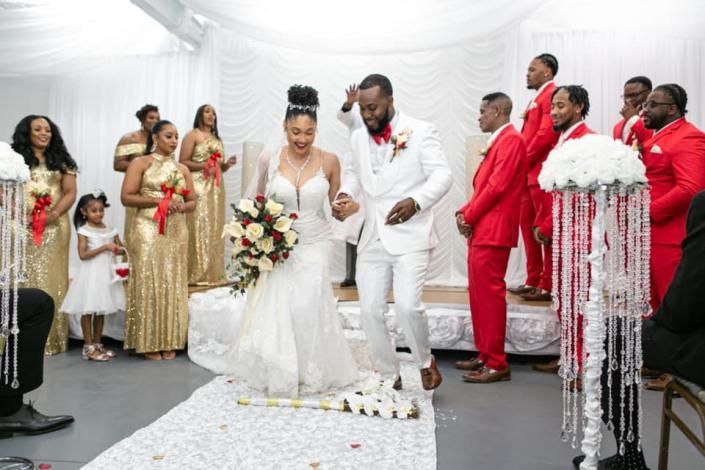 Wedding couple in white, walking down an aisle with guests under a white draped canopy.