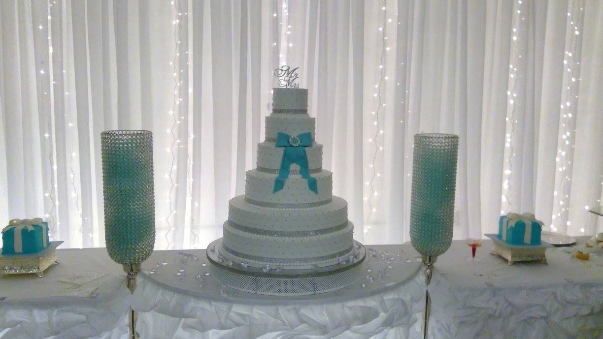 White tiered cake with teal ribbon topper, flanked by tall teal candle holders on a decorated table.