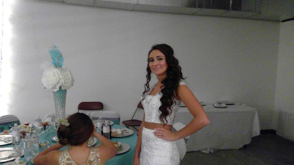 Bride in a white dress stands indoors beside a decorated table with a seated guest.