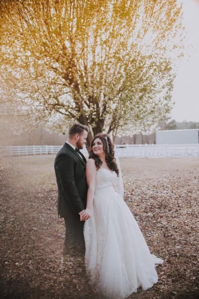 Bride and groom standing beneath a golden autumn tree in a field, holding hands and smiling