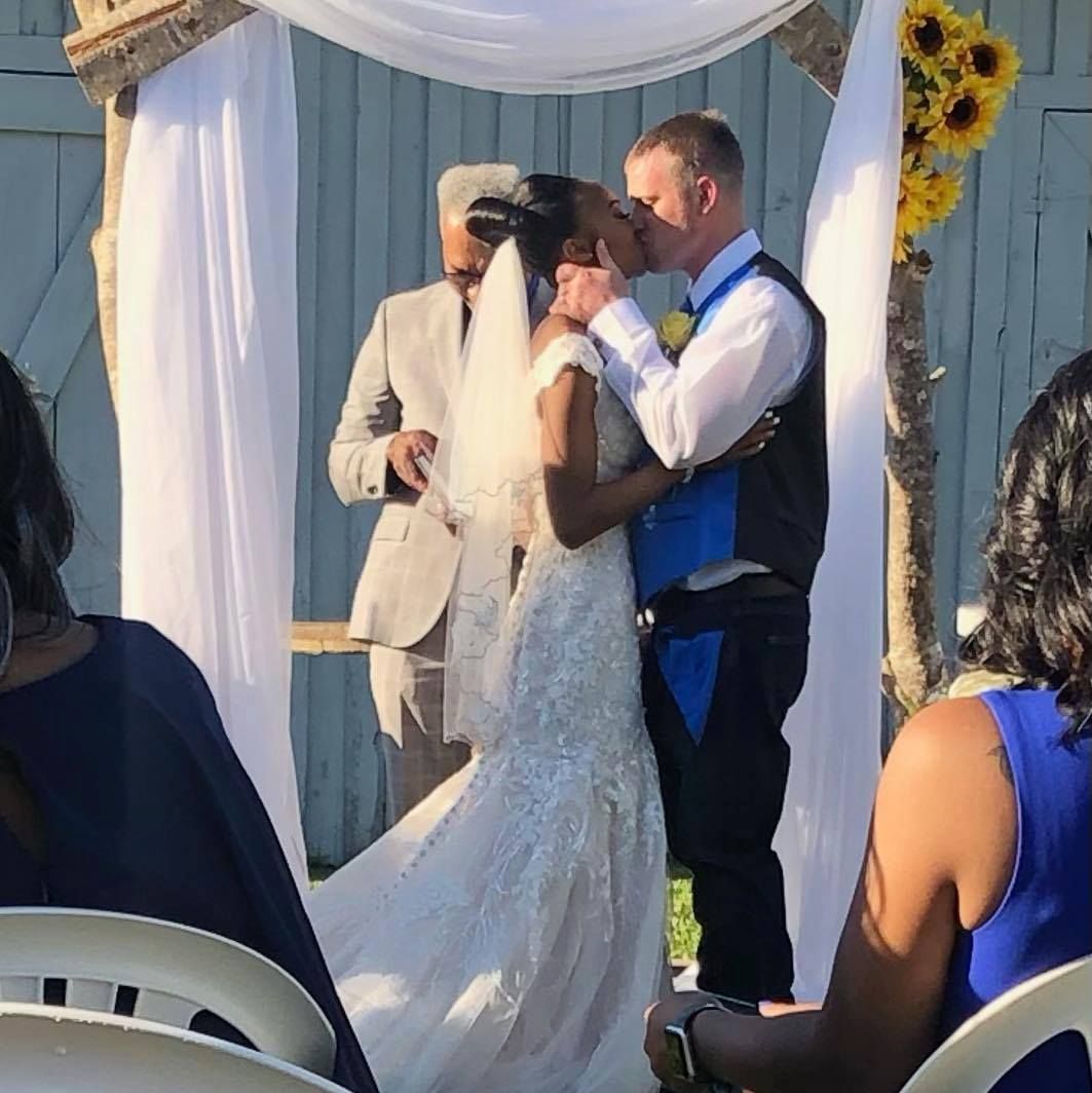 Bride and groom share a kiss under a white wedding arch, with a sunflower accent and seated guests nearby.