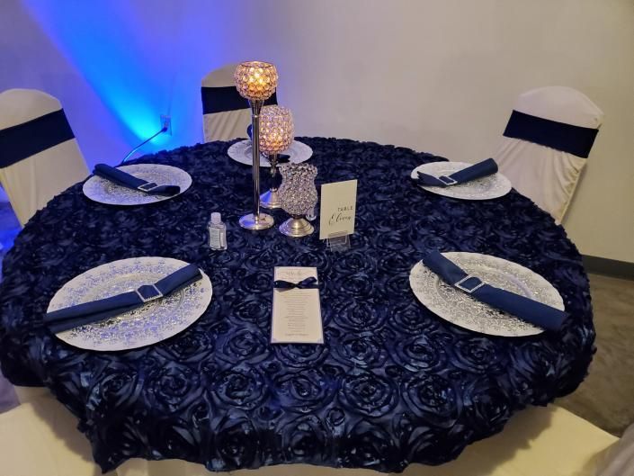 Decorated round dining table with blue tablecloth, plates, napkins, glasses, and candle centerpiece