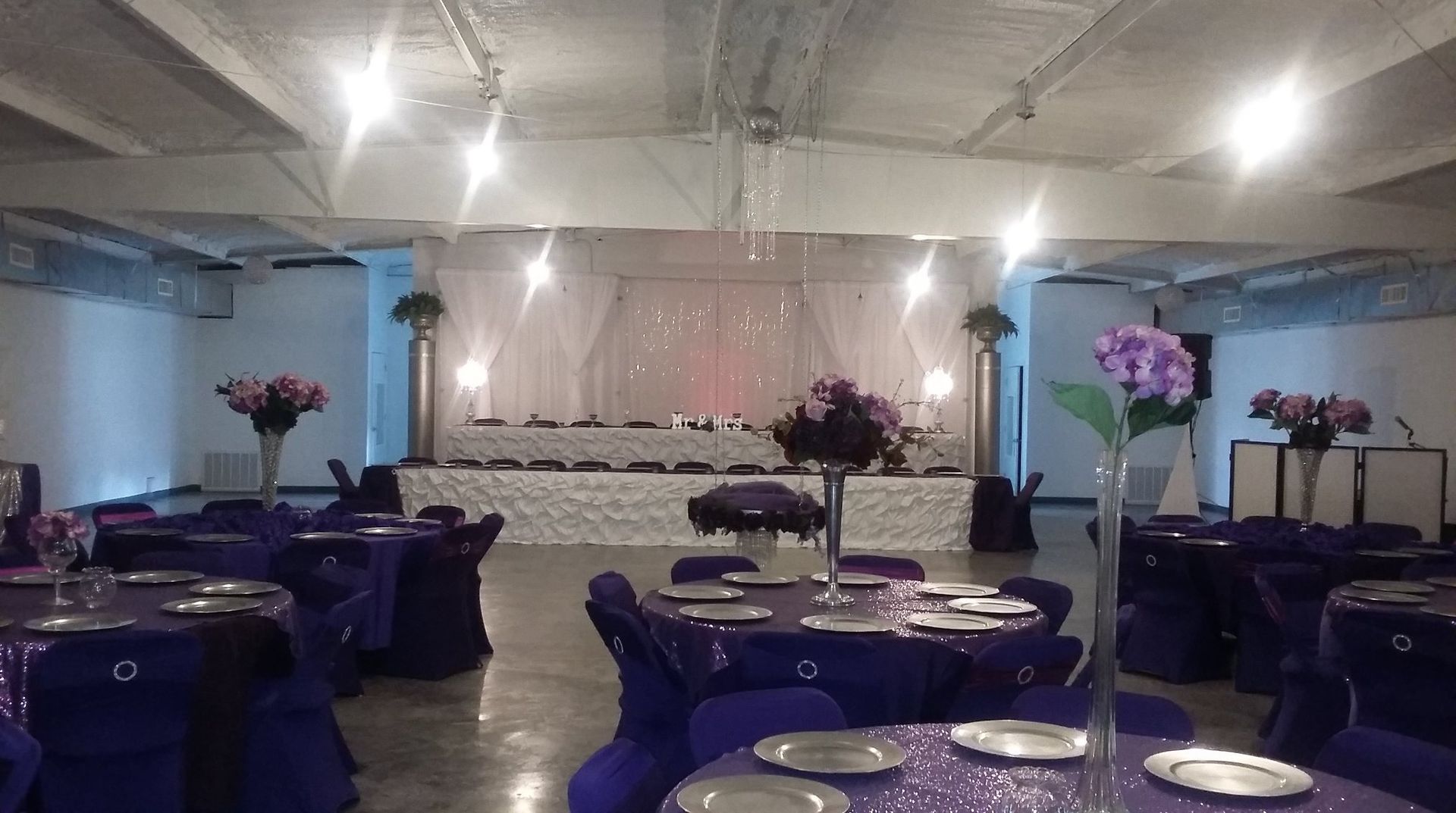 Elegant banquet hall with purple table settings, floral centerpieces, and a decorated stage at the back
