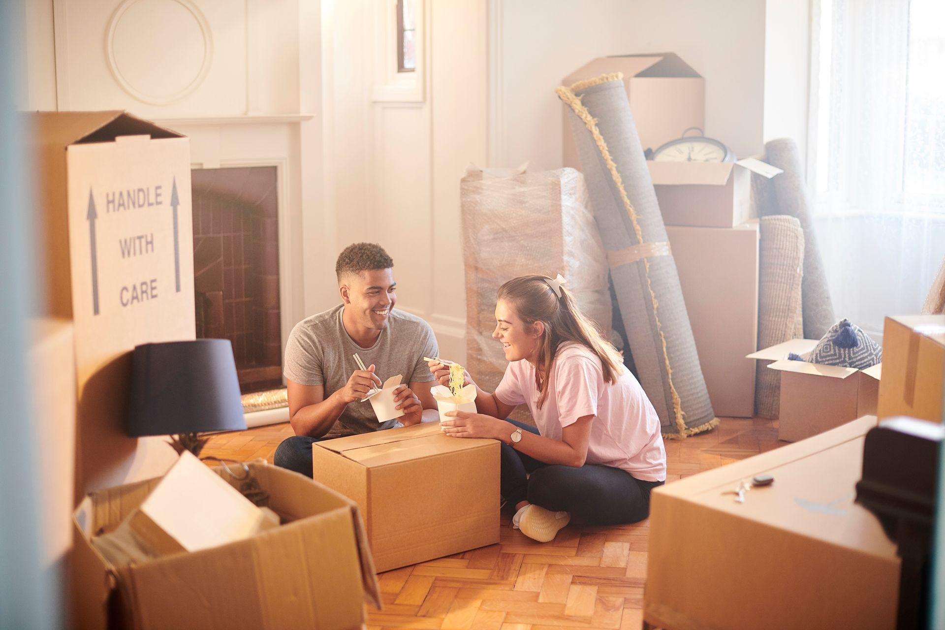 Couple packing moving boxes in a sunlit room with stacked furniture and a lamp