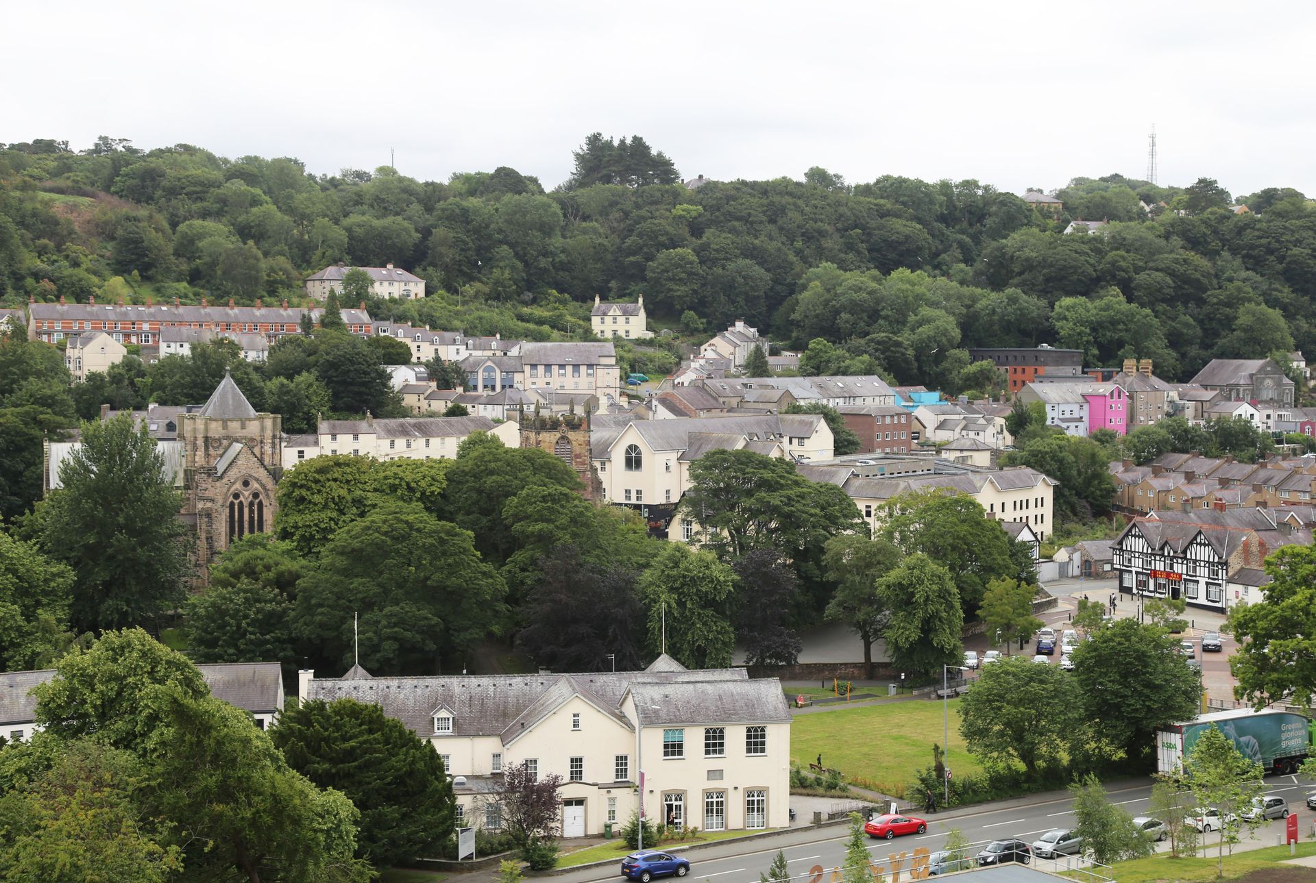 Hillside image of the city of Bangor, North Wales with stone houses, lush trees, and a road in the foreground under an overcast sky