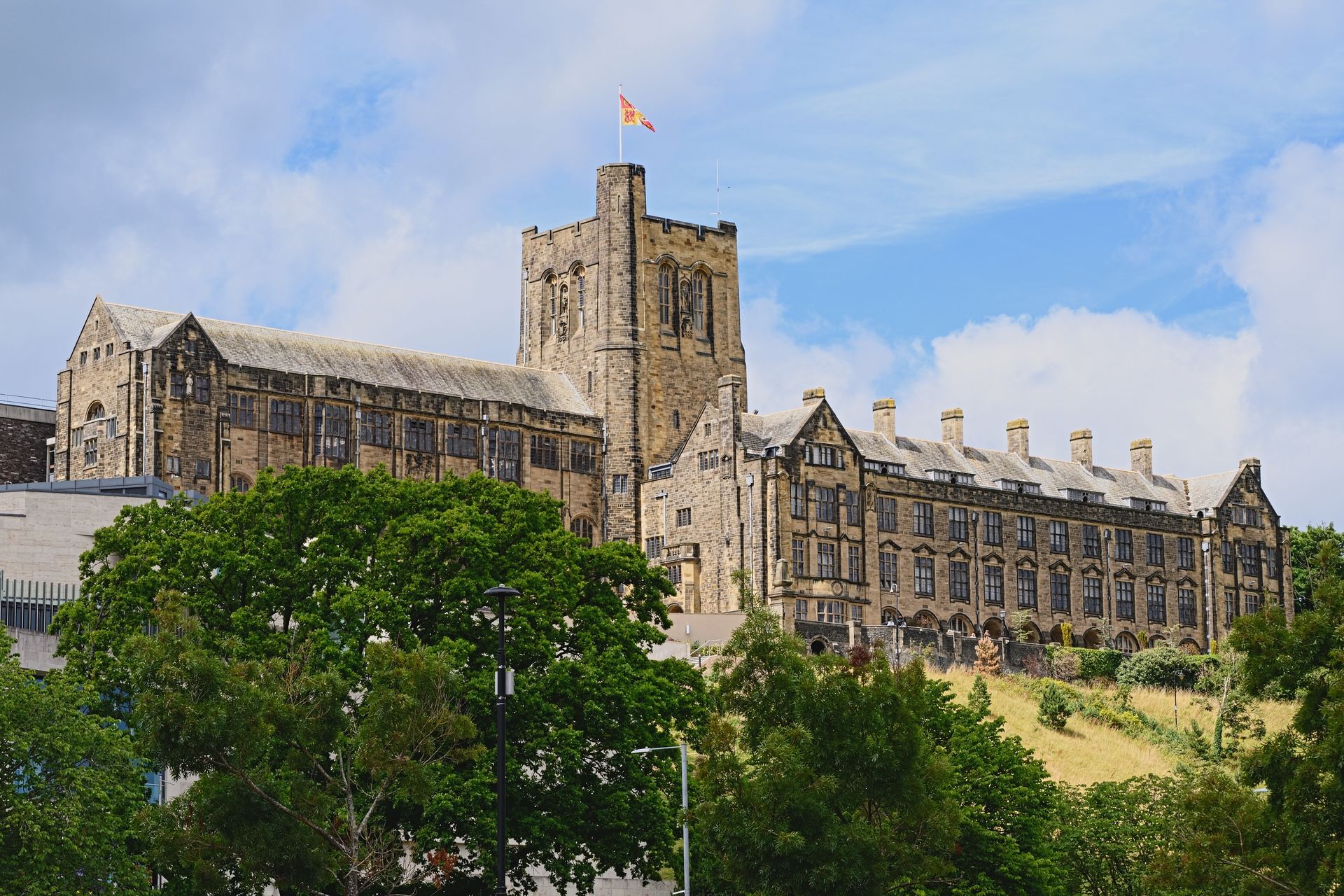 Stone front facade of Bangor University, North Wales on a hill with a central tower, blue sky, and trees in the foreground