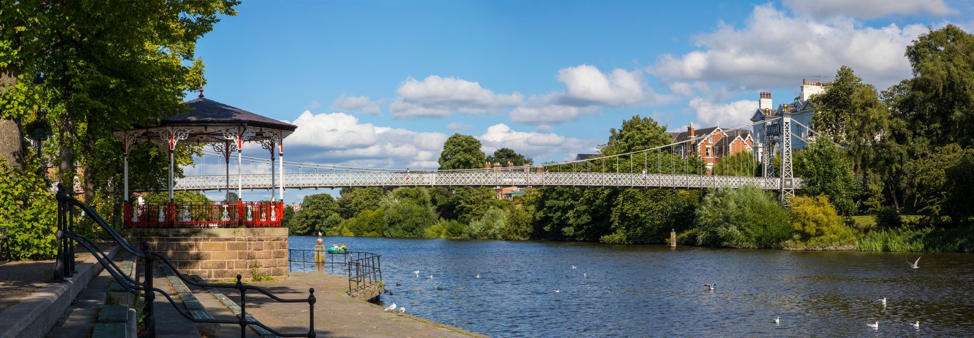 Steel suspension bridge over the River Dee in Chester with trees, and a gazebo under a partly cloudy blue sky