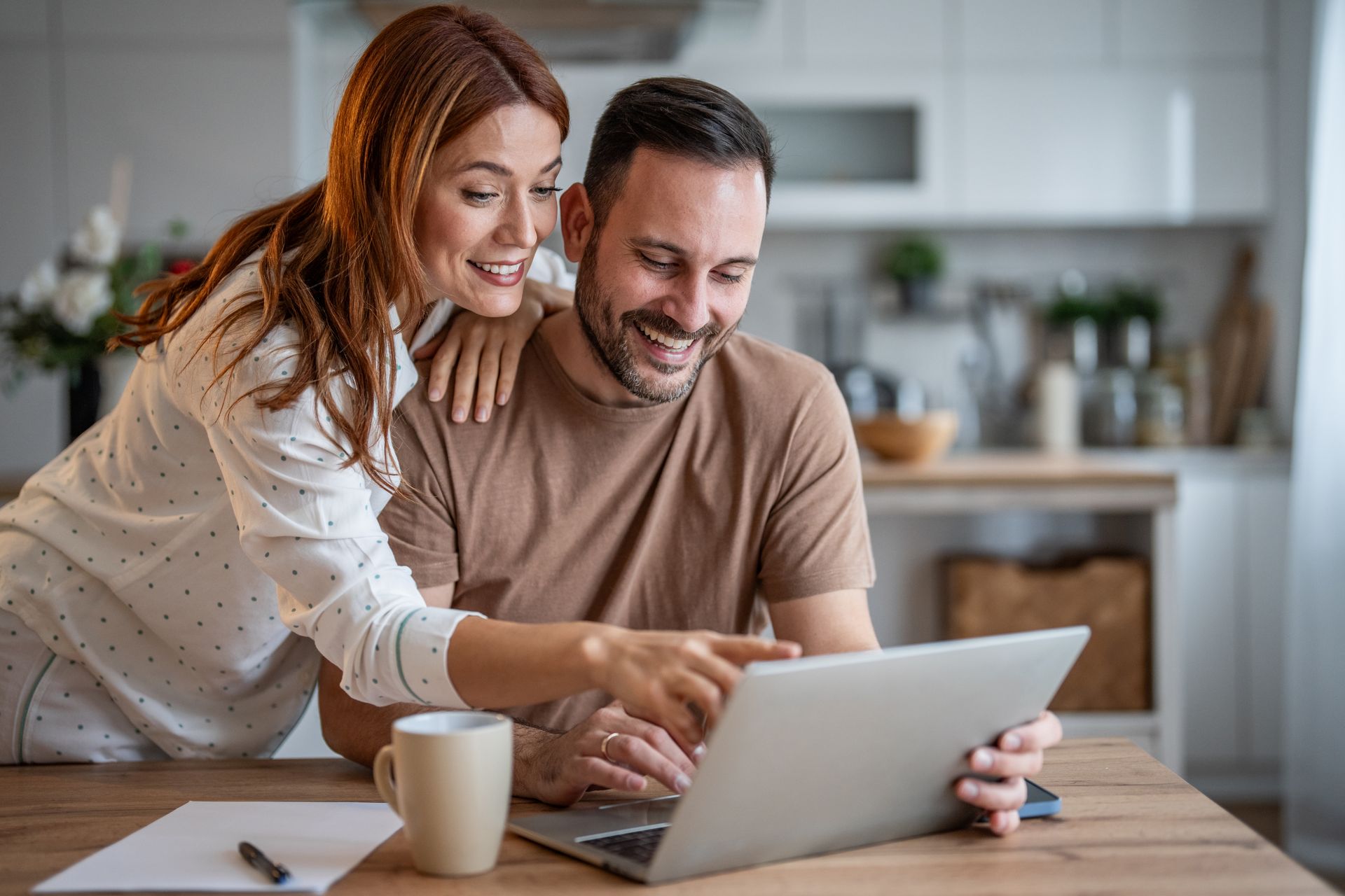 Two people smiling and looking at a laptop in a bright kitchen getting a FREE Quote online, with a coffee mug on the table.