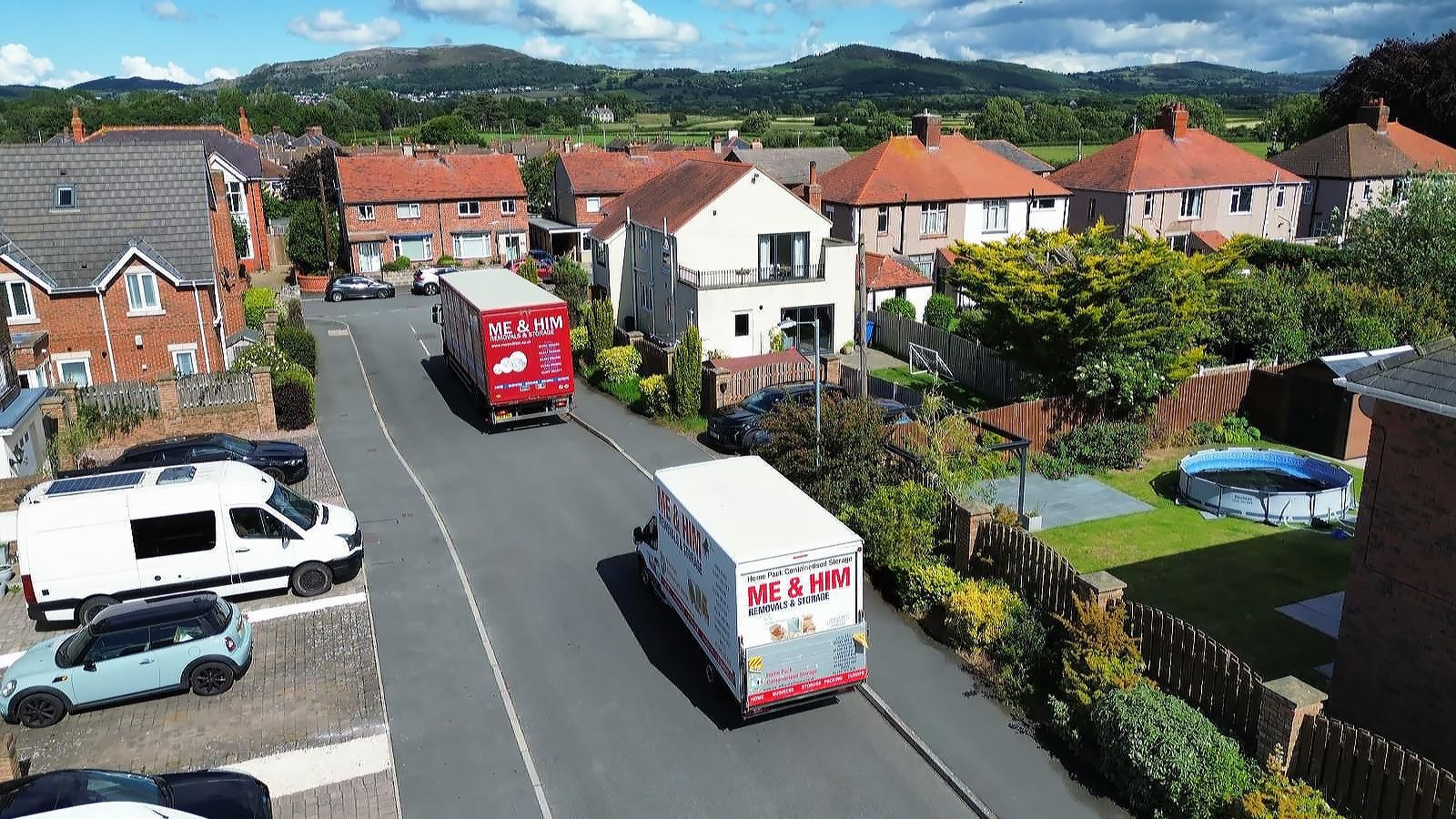 Aerial view of a suburban street with parked vans, red-roofed houses, and hills in the background and Me & Him Removals & Storage vehicles driving through.