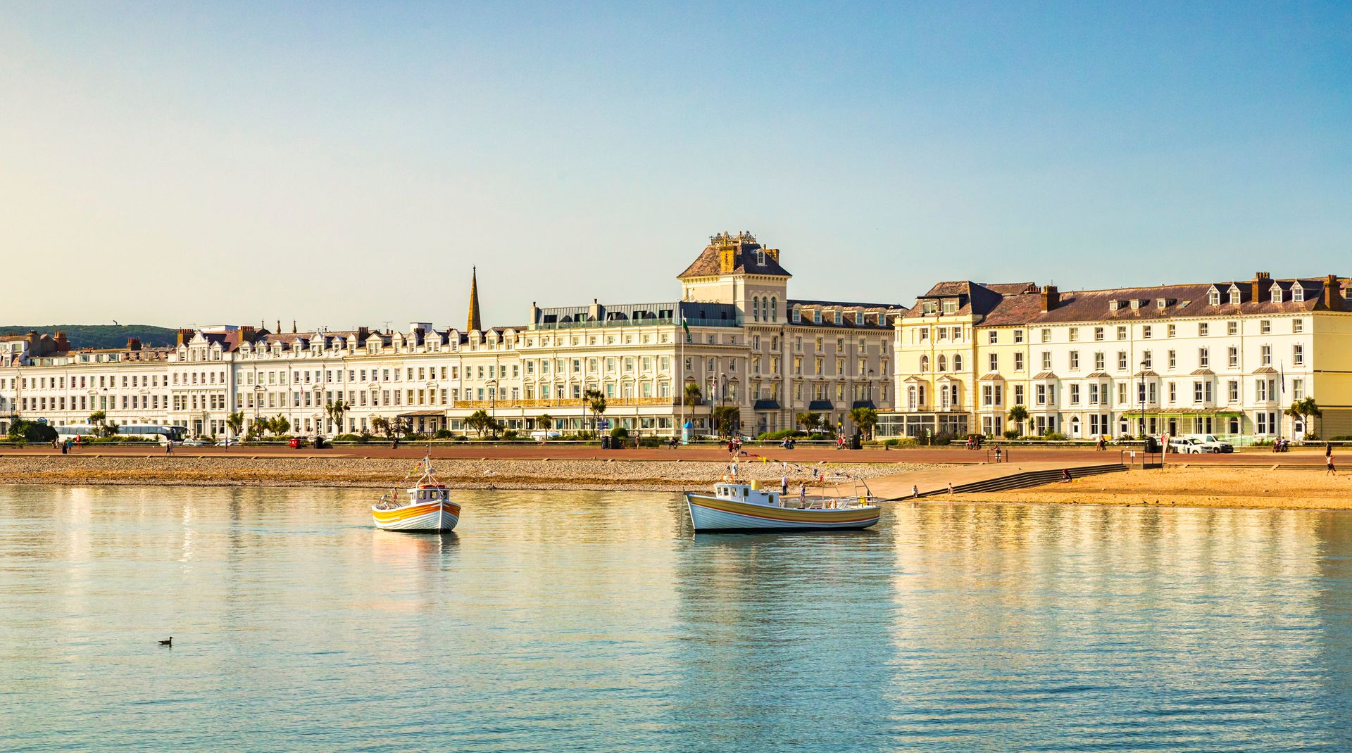 Sunny waterfront promenade in Llandudno, North Wales with white Victorian buildings, boats, and calm blue water under a clear sky