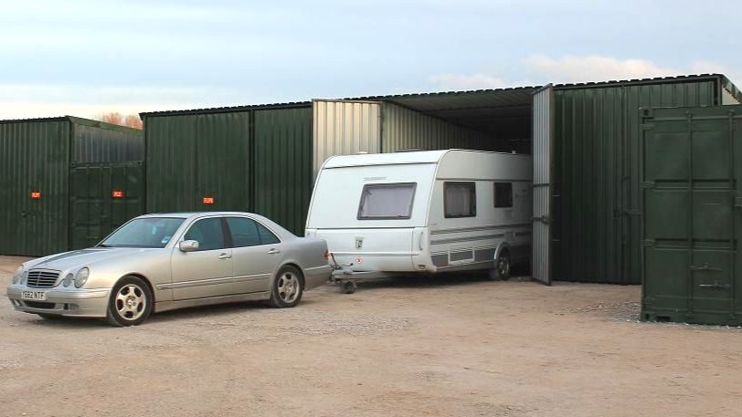 Silver Mercedes car towing a white caravan from a green secure storage unit onto a gravel compound