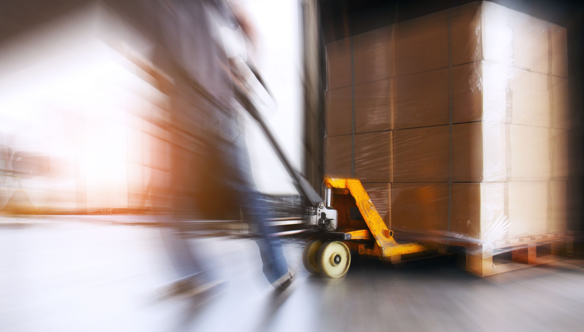 Me & Him Removals & Storage moving stacked cardboard boxes on a pallet in a warehouse aisle with motion blur.