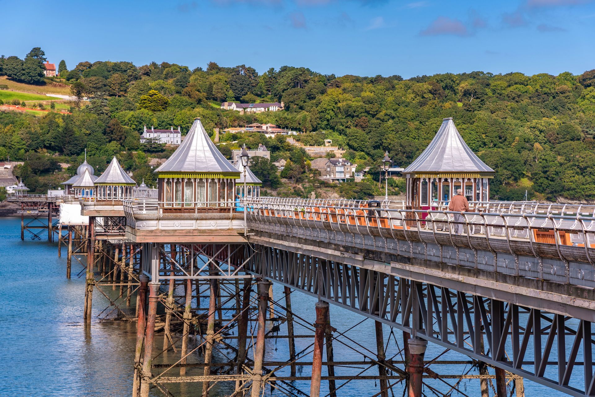 Wooden pier in Bangor, North Wales  with white pavilions over blue water, stretching toward a green hillside coastline