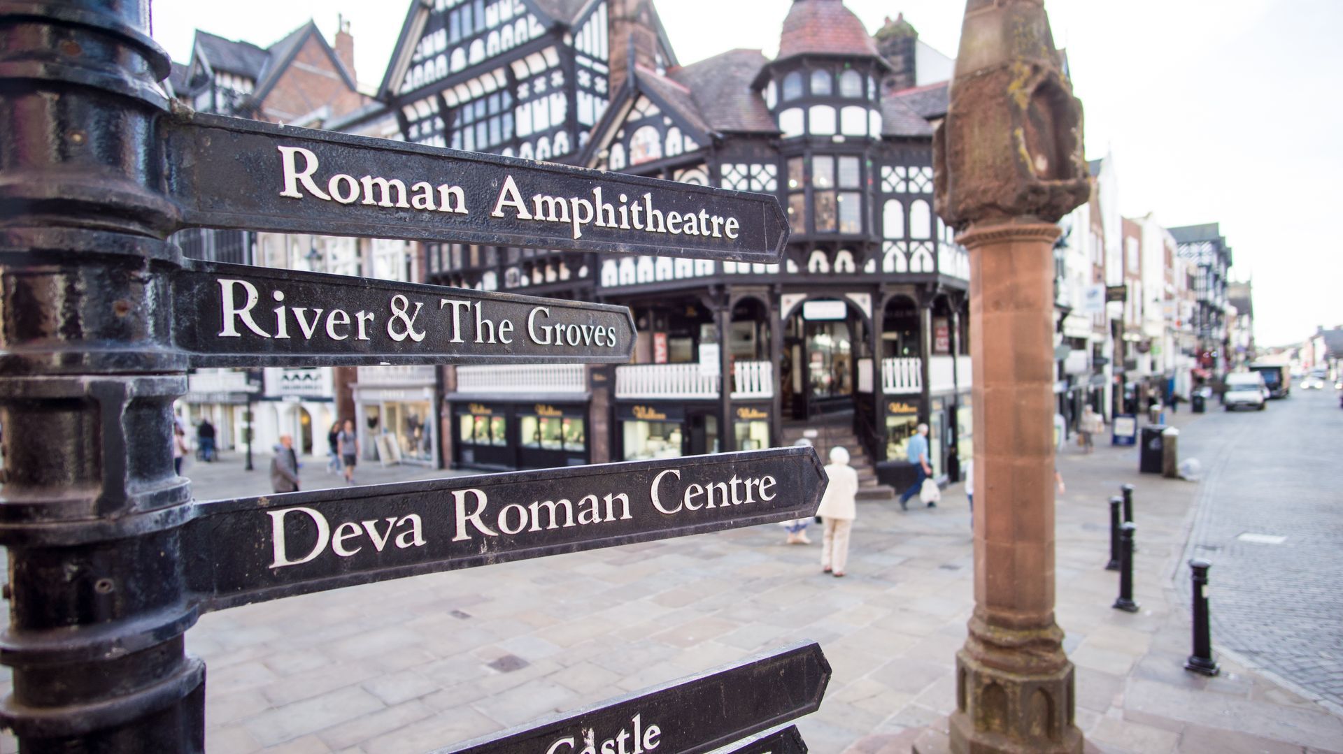 Tourist signs for Roman Amphitheatre and Deva Roman Centre in Chester city centre, with a stone lamp post nearby