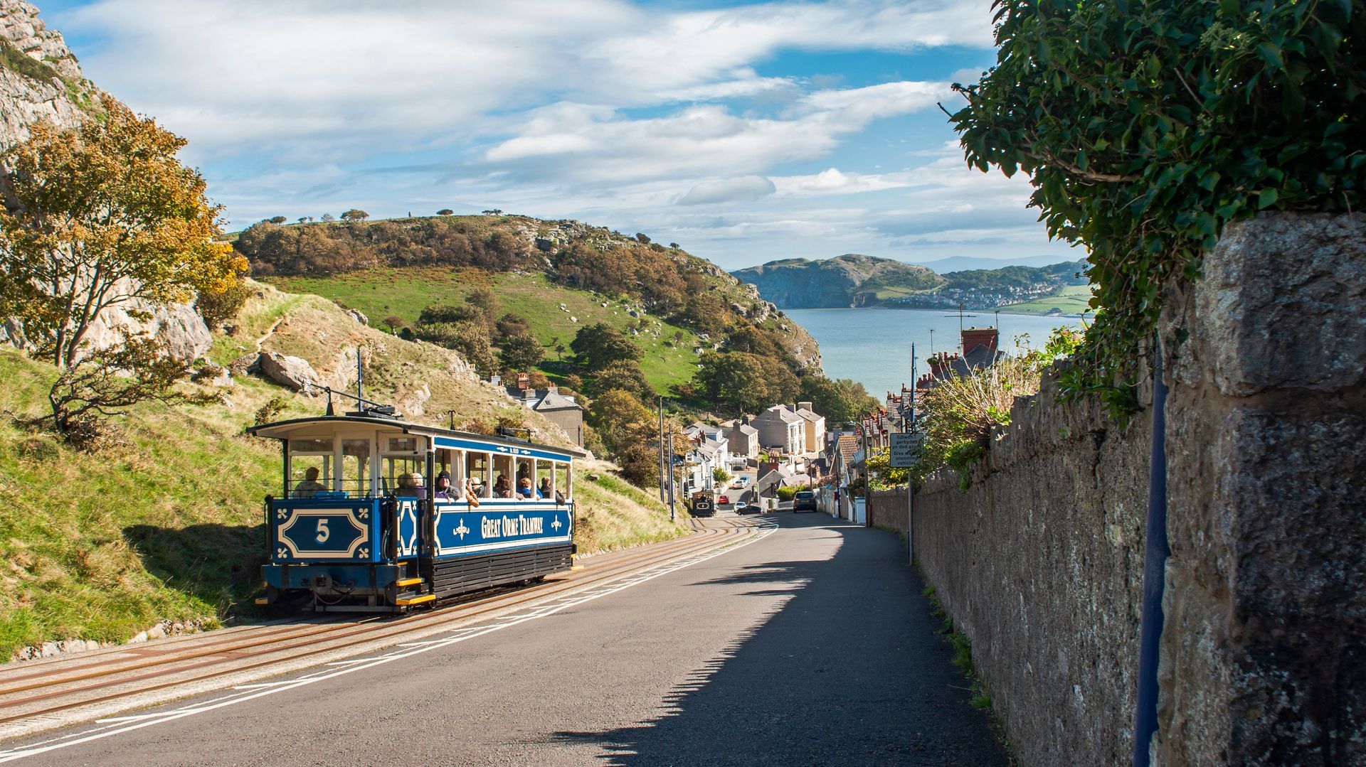 Blue tourist tram on a seaside road in Llandudno, North Wales with hills, stone wall, and harbour in the distance