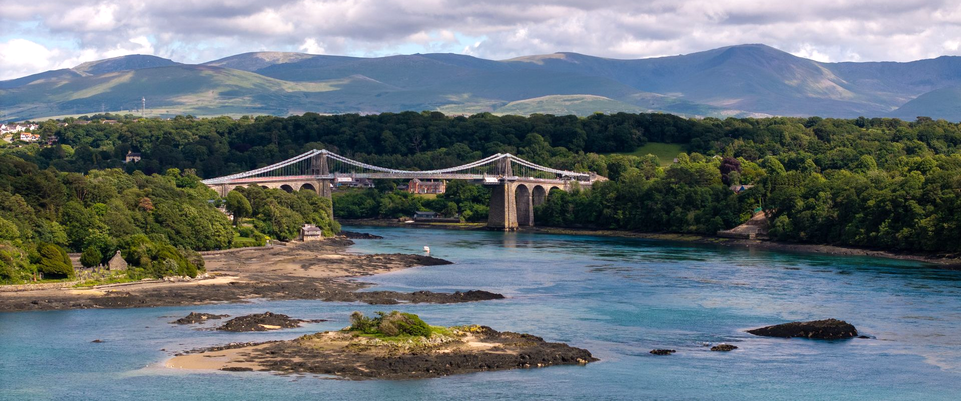Menai suspension bridge over the blue Menai Straits with green trees and distant Eyri (Snowdonia National Park) mountains under cloudy skies in the distance.
