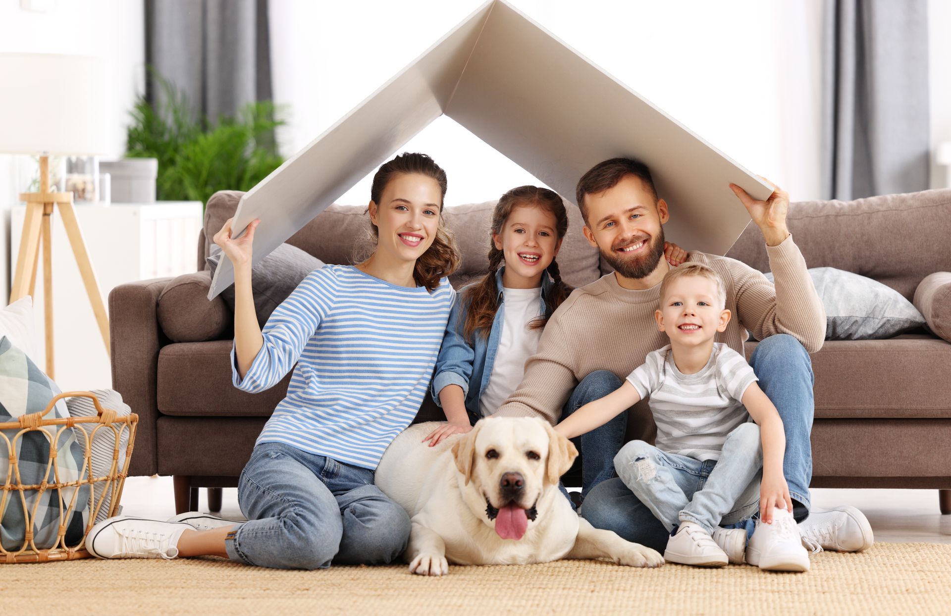 Family sitting on the floor with a dog inside a living room, smiling and holding a cardboard roof playhouse.