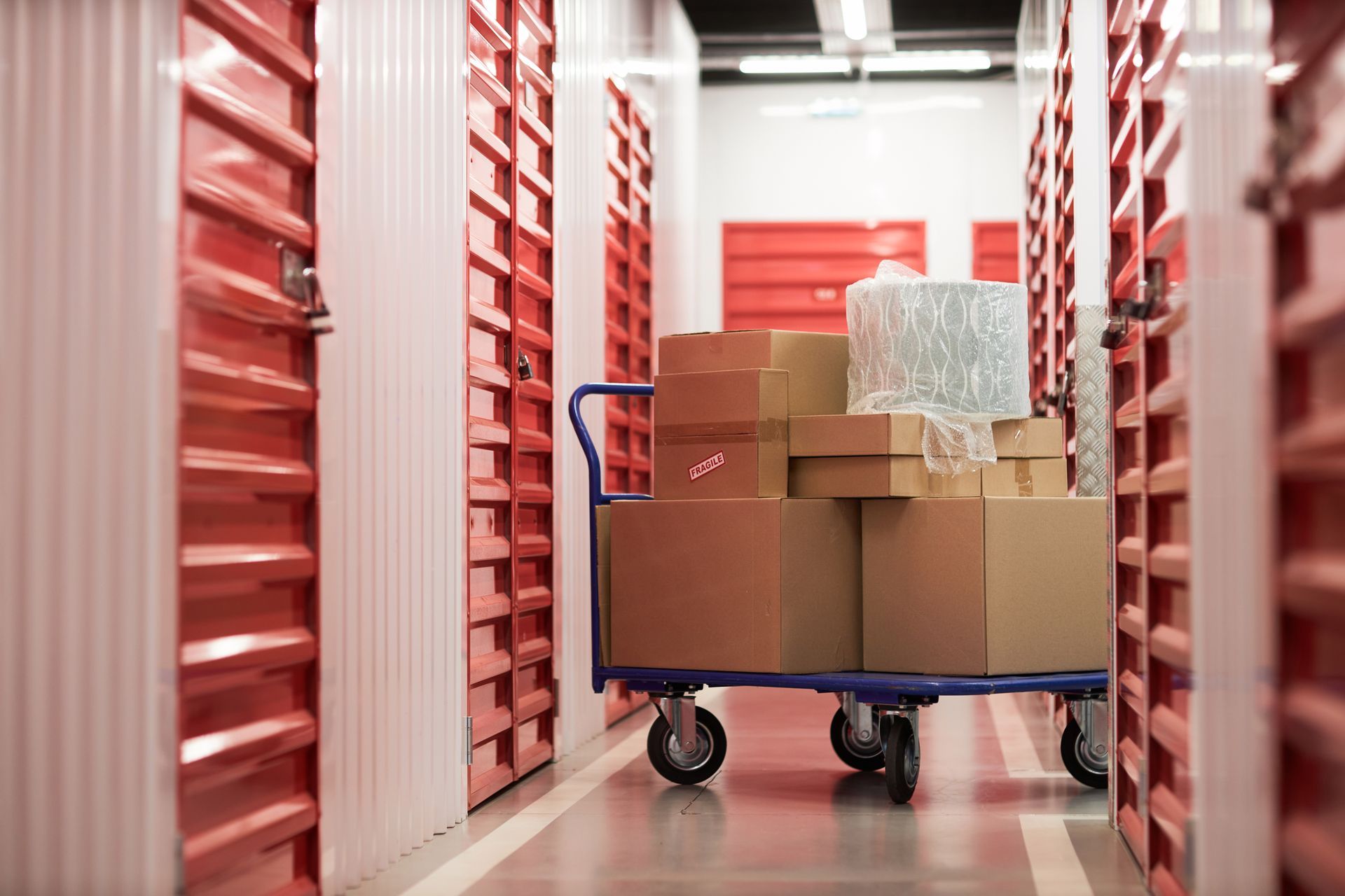 A storage facility hallway with red unit doors and a white floor under bright ceiling lights.