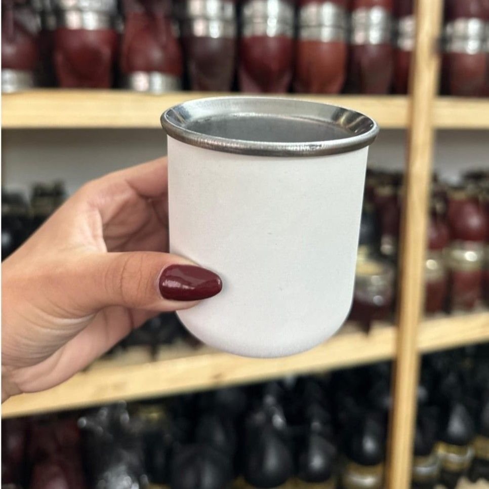 Una mano sostiene una pequeña taza blanca con borde plateado frente a estantes llenos de frascos y botellas.