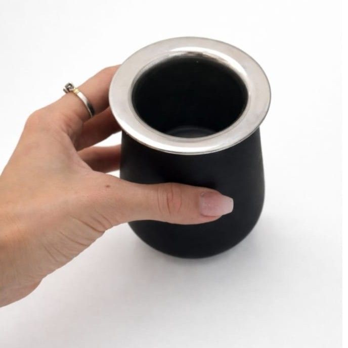 Mano sosteniendo un vaso térmico negro con borde plateado sobre un fondo blanco.