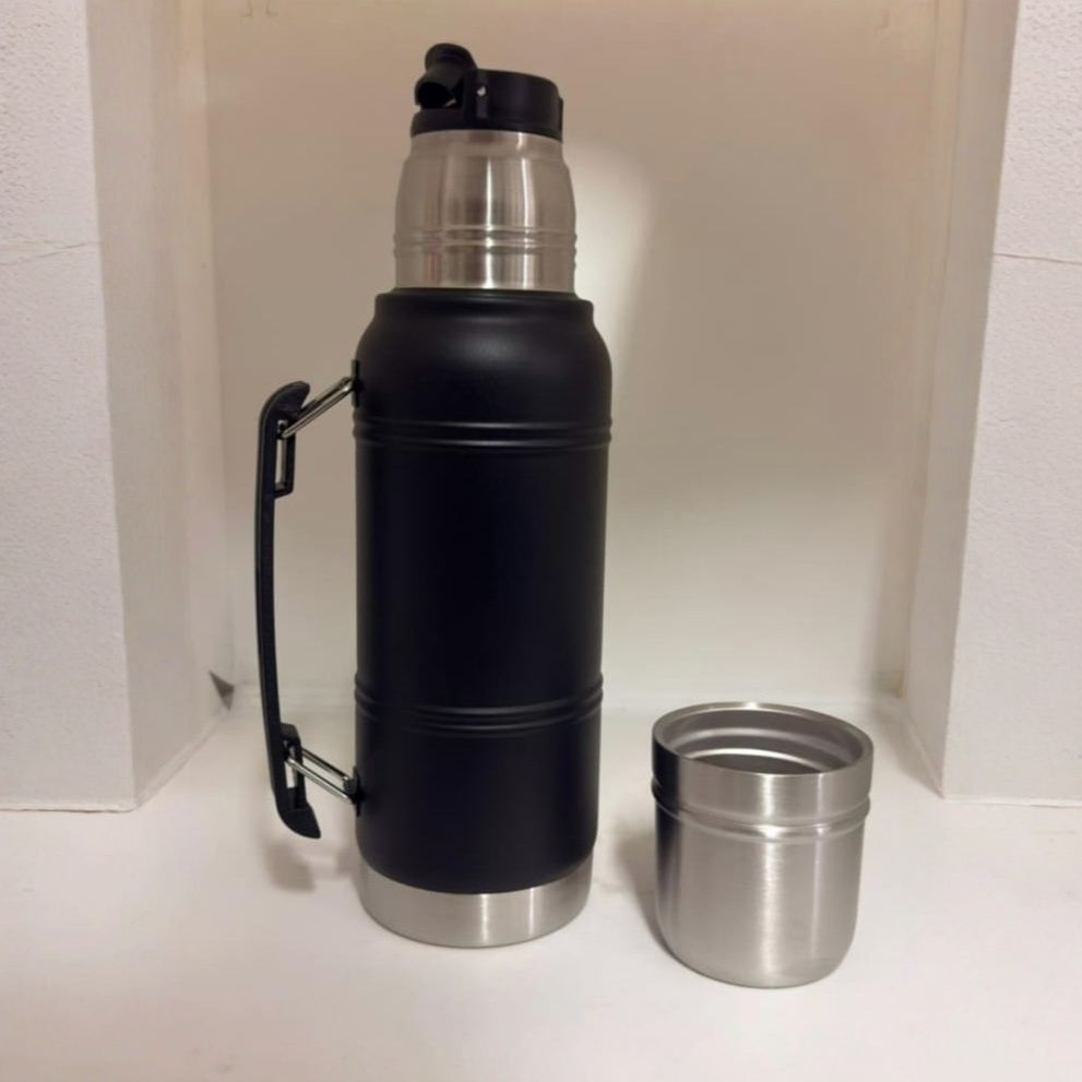 Botella de agua isotérmica negra con asa junto a una taza de acero inoxidable sobre una superficie blanca.