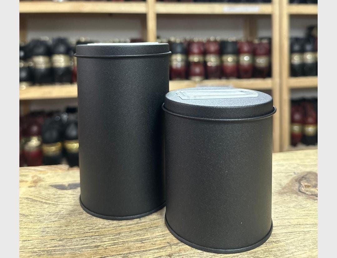 Dos recipientes cilíndricos de color negro mate sobre una mesa de madera, con estantes de frascos difuminados al fondo.