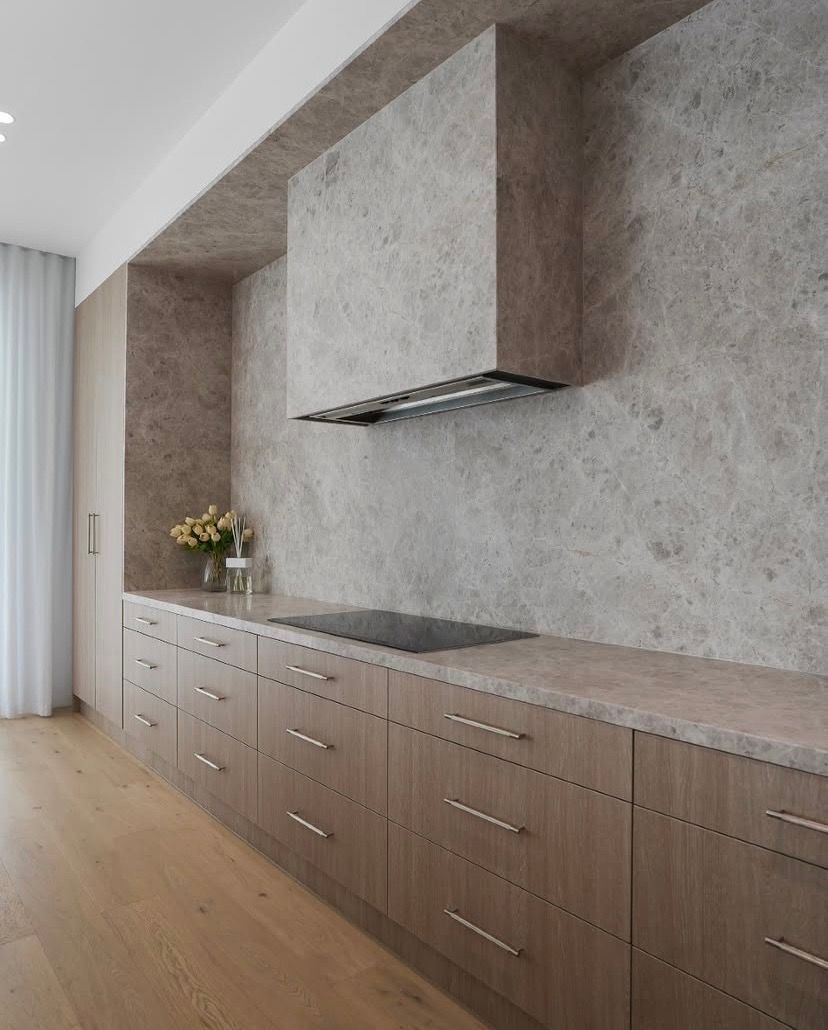 A modern kitchen featuring wooden cabinets, a light stone countertop, and a matching textured backsplash and range hood.
