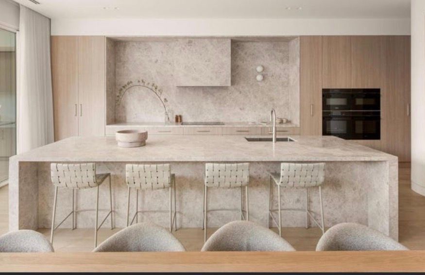 Modern, neutral-toned kitchen featuring a large stone island with four woven bar stools, wood cabinetry, and an oven.