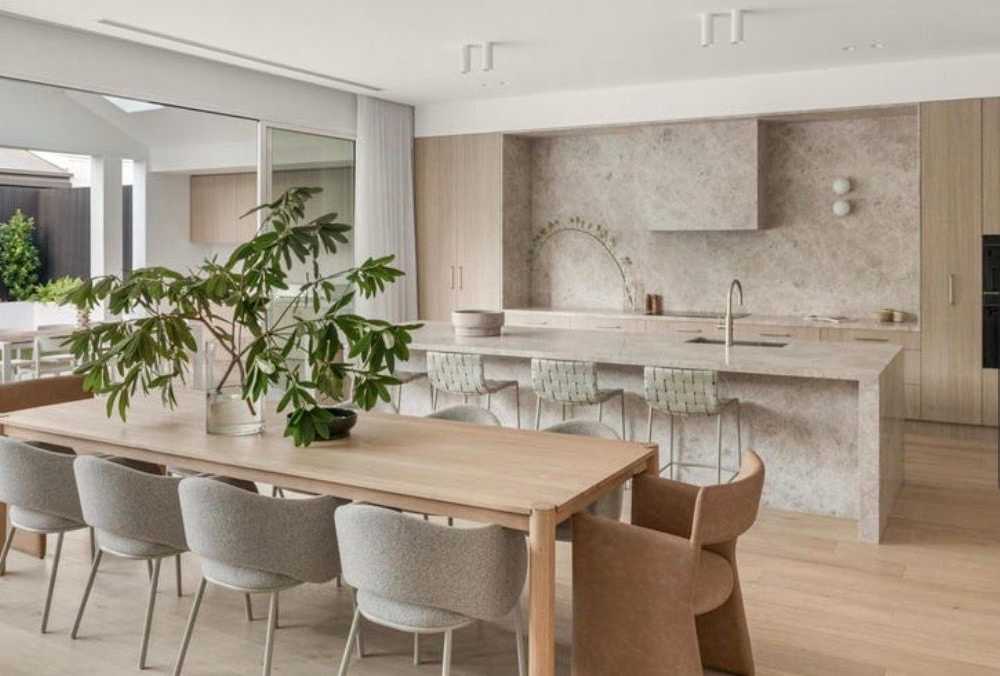 A bright, modern open-plan kitchen and dining area with a long wooden table, light stone island, and neutral decor.