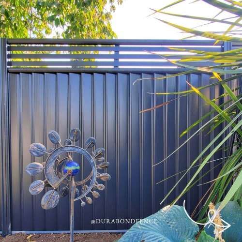 Modern backyard with dark vertical fence, blue decorative wind spinner, and tropical plants in bright sunlight