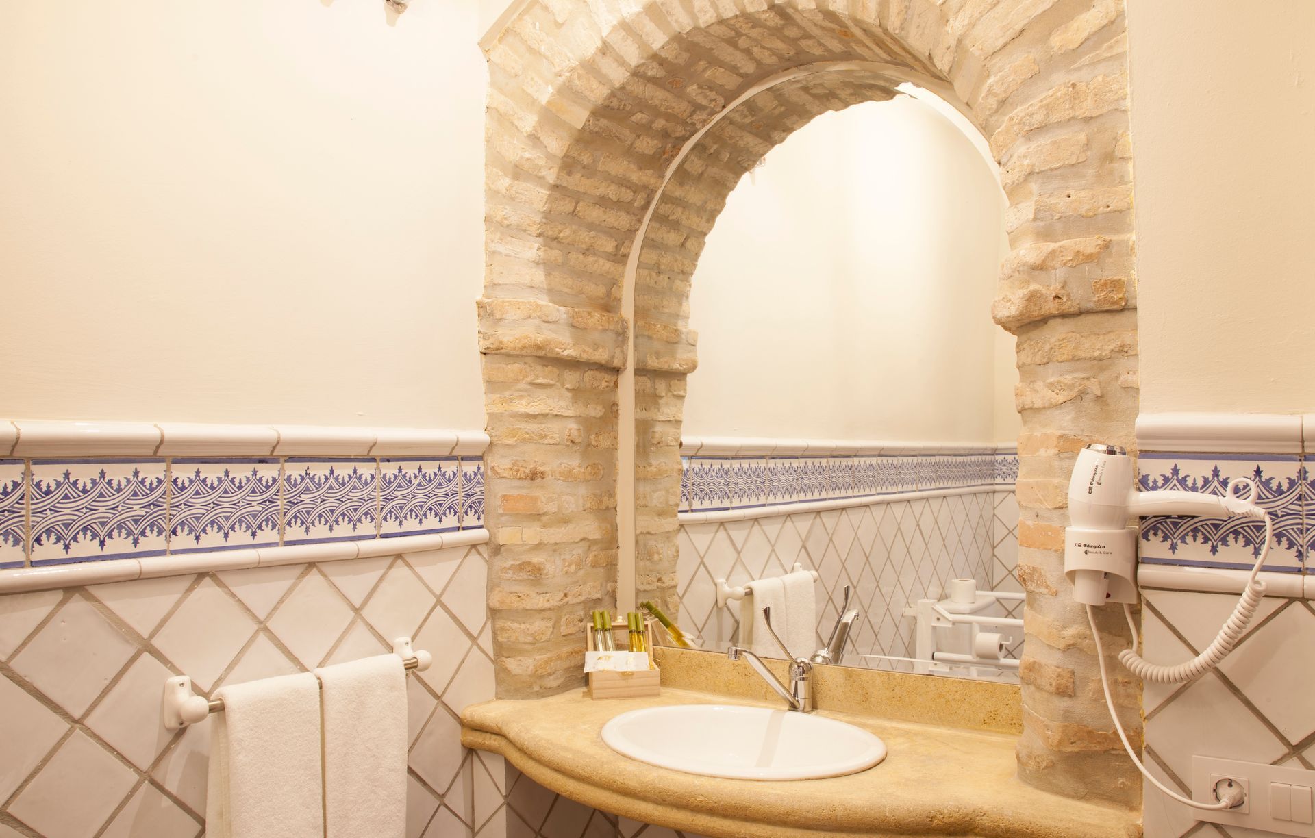 Bathroom sink with stone arch mirror, beige tiles, and decorative blue border around the walls
