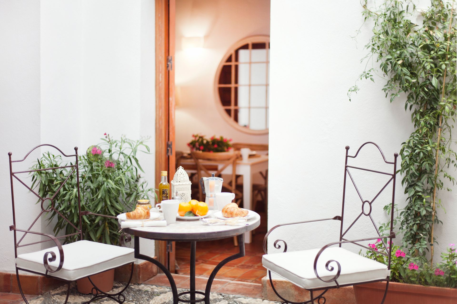 Outdoor patio with two white chairs, a round table set with drinks, and potted plants by an open doorway