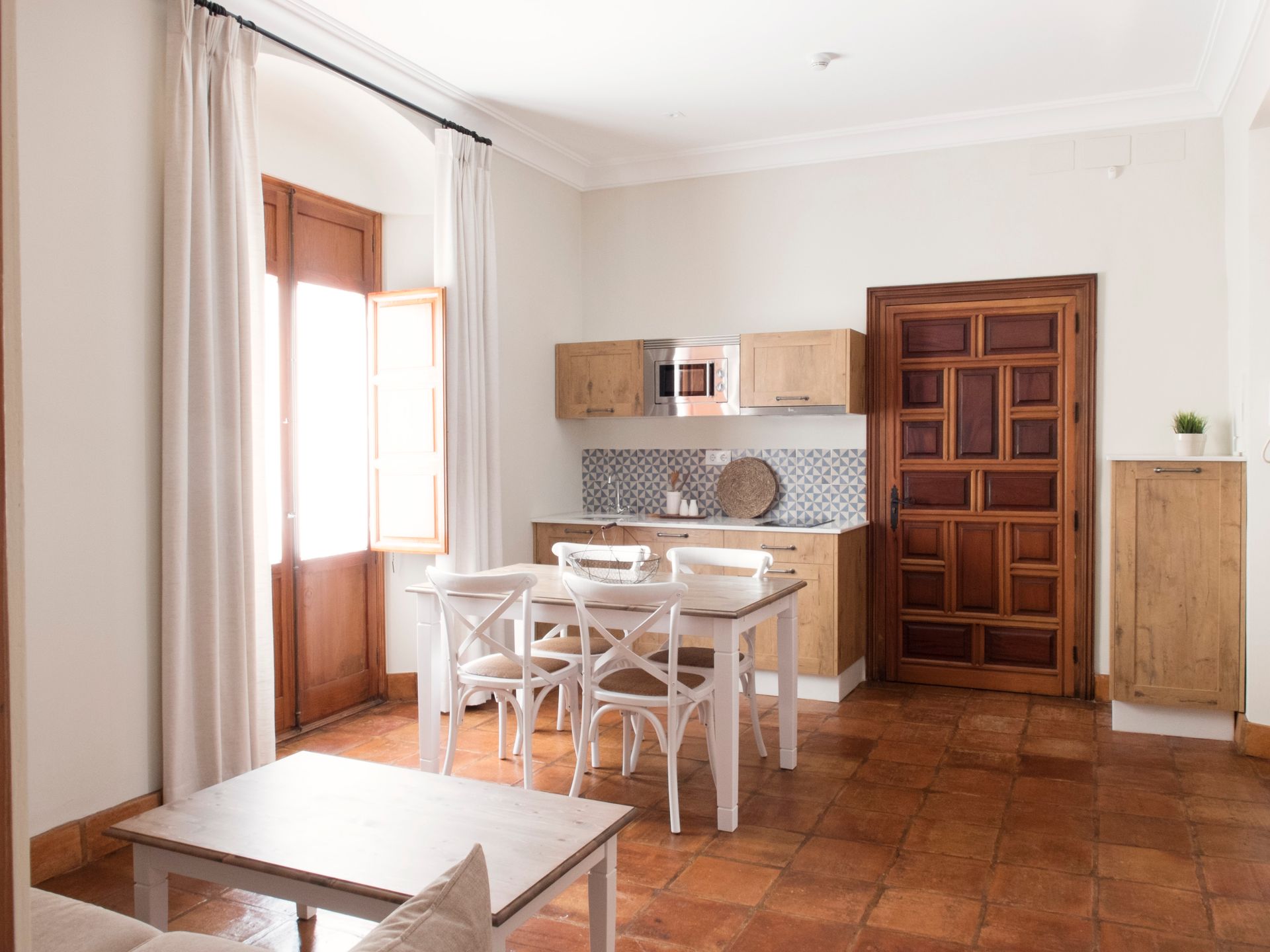 Bright dining area with a small wooden table, white chairs, tiled floor, and a wooden door