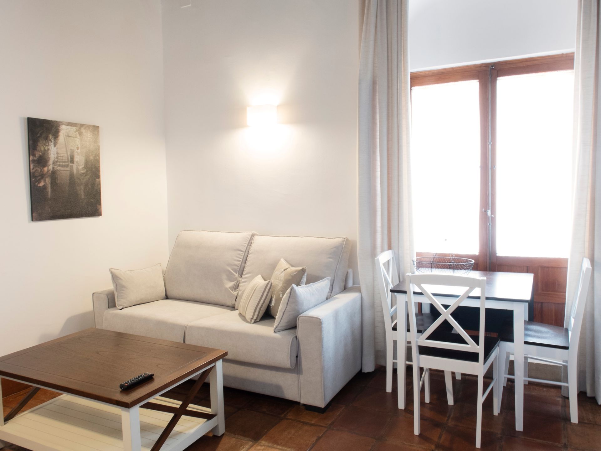 Bright living room with a beige sofa, wooden coffee table, white dining set, and large window with curtains.