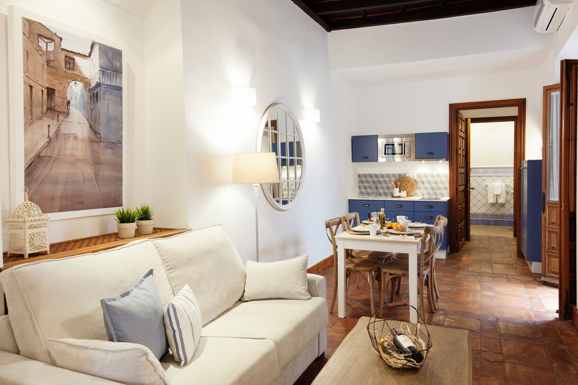 Cozy living room with white sofa, dining table, blue kitchen, and warm terracotta tile flooring