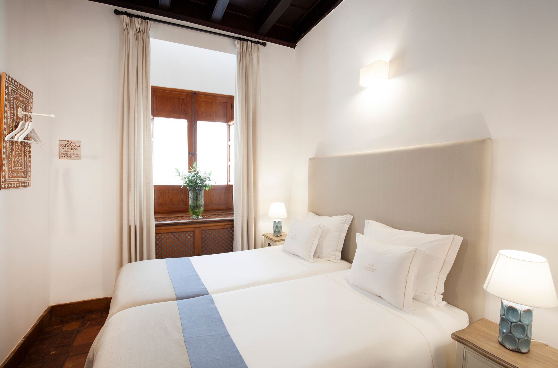 Bright bedroom with twin beds, white walls, wooden floor, and a window with curtains and a potted plant.