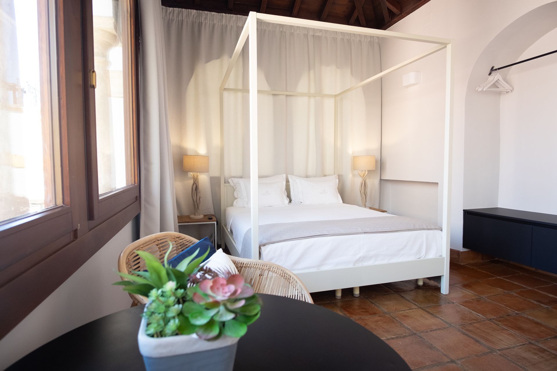 Bright bedroom with white canopy bed, wooden floor, and table with potted flowers by the window