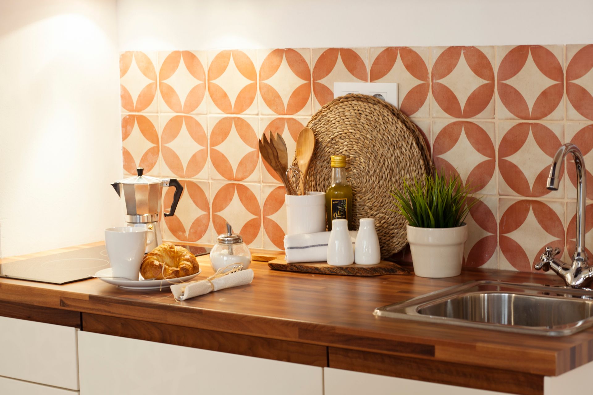 Modern kitchen counter with coffee setup, wooden cutting board, patterned orange tile backsplash, and sink
