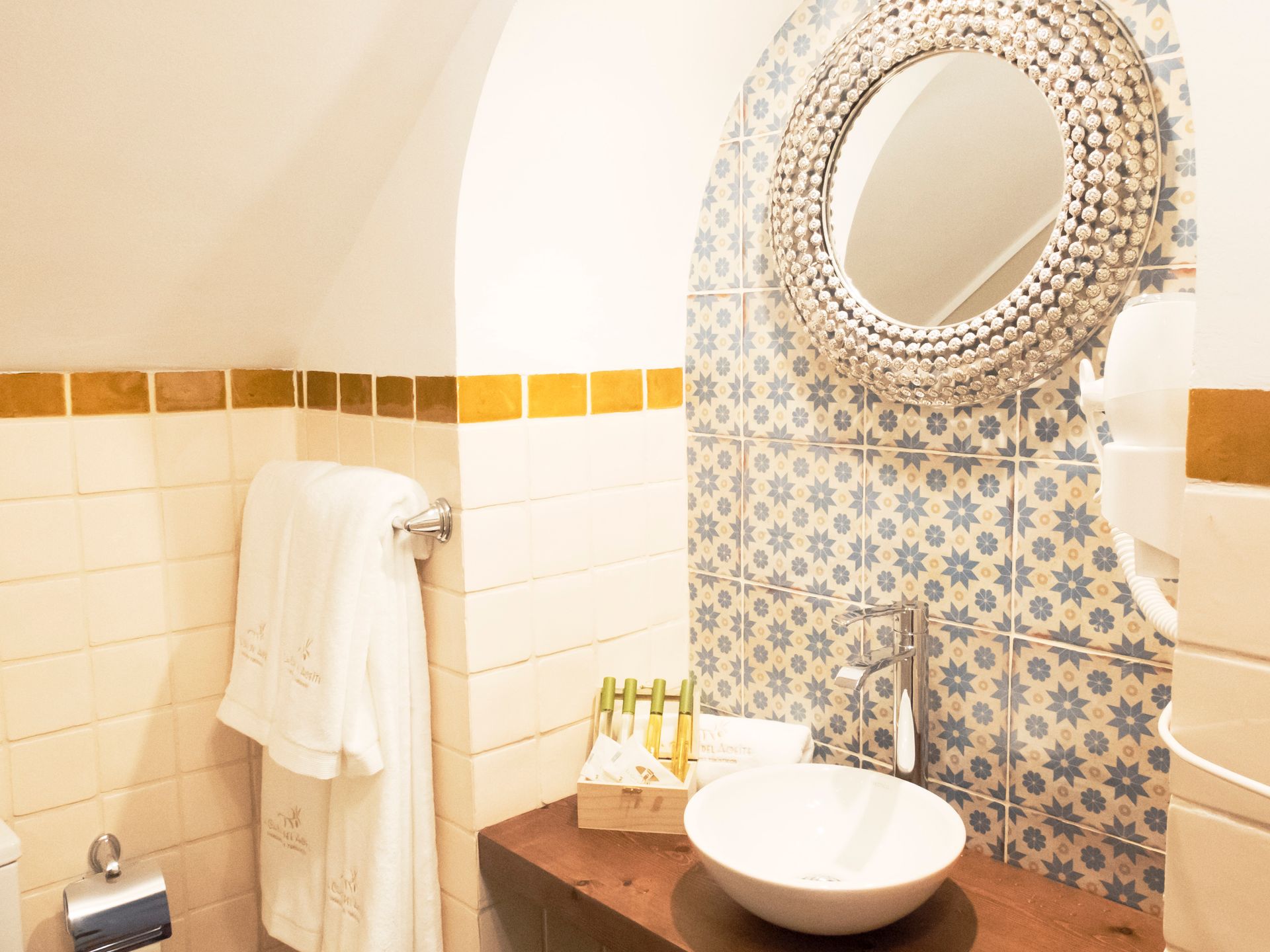 Small bathroom vanity with round mirror, white sink, patterned tile, and towel on wall rack