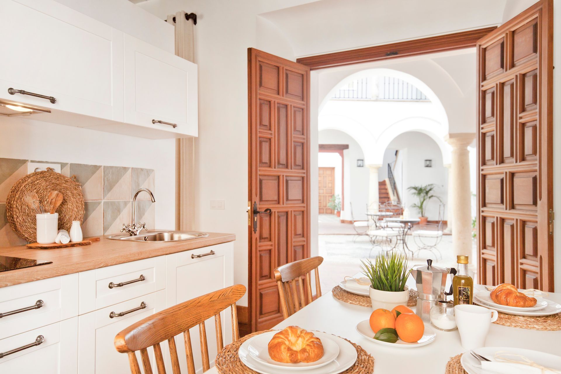 Bright kitchen with wooden double doors open to a sunny courtyard and a table set with fruit and pastries