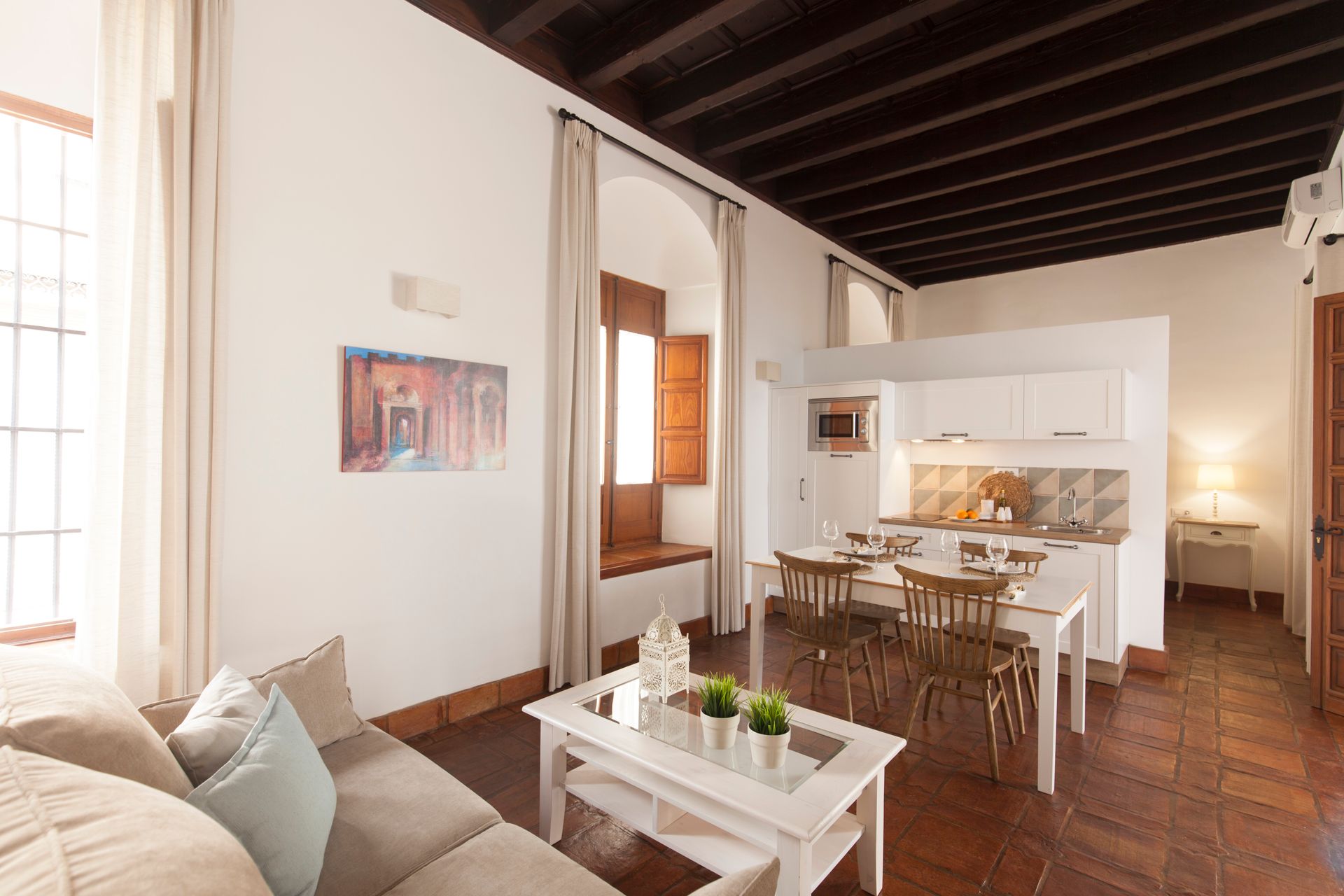 Bright living-dining room with white furniture, exposed dark beams, and a kitchen in the background